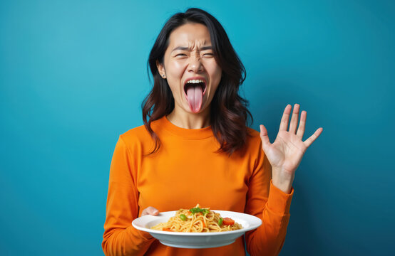 Asian woman in orange shirt displays disgust for instant noodles, sticking out tongue with grimace. Holds plate of food, gesturing negatively. Vibrant blue background emphasizes strong negative