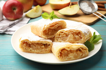Pieces of tasty apple strudel with powdered sugar and mint on light blue wooden table, closeup