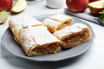 Pieces of tasty apple strudel with powdered sugar and fruits on white marble table, closeup