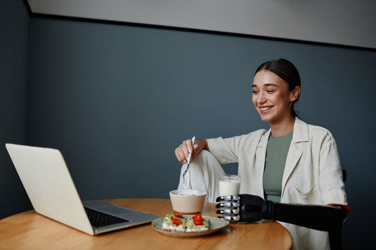 Happy woman with black bionic arm eating healthy breakfast and watching video on laptop at table