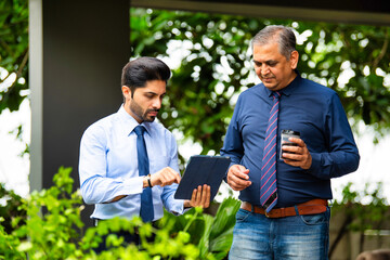 Indian corporate professionals walk with tablet, engaged in discussion outside office building