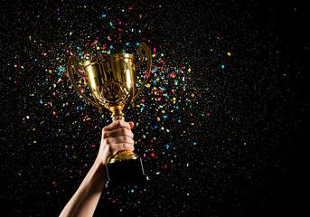 Celebrating a triumphant victory with a golden winner's trophy cup raised high amidst a burst of colorful confetti against a dark background.