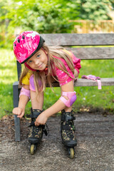 Happy little girl in protective helmet and knee and elbow pads sitting on a bench and putting on...
