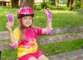 Happy little girl puts on a protective helmet and pink elbow pads for safe rollerblading or...