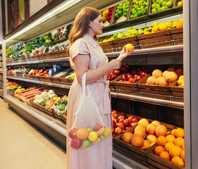 Young woman with string bag in grocery store. Female holding a lemon while standing at shelves in a grocery store.