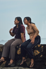 A calm moment as two teenage girls sit side by side near the beach, eyes drawn toward something in the distance, enjoying the coastal view in silence.