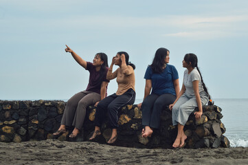The girls share smiles and conversations by the beach; one of them points toward the distance, likely at something interesting across the water.