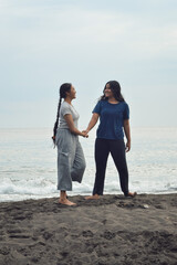 Two girls walk side by side, one reaching out, both sharing a light moment while approaching a friend, with morning waves curling behind them.