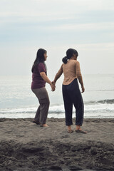 While walking hand in hand, the girls pause and look curiously at something off-frame, standing on the sunlit sand near the calm sea.