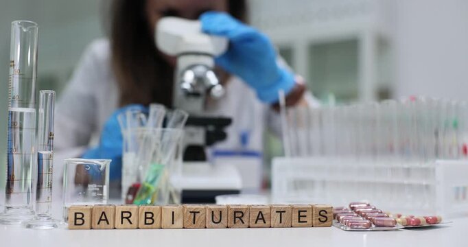 Wooden cubes spell Barbiturates showing dangerous substances topic. Scientist works at table in modern laboratory observing samples through microscope