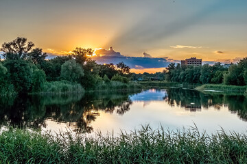 Obraz premium Dramatic sunset over Khorol River with golden reflections, lush vegetation, and urban buildings forming a picturesque evening landscape in Myrhorod, Ukraine