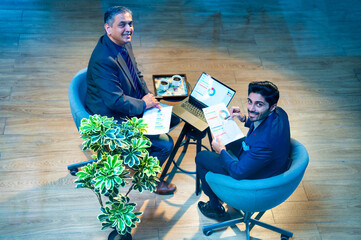 Indian corporate partners collaborate while seated at desk using laptop in top-angle office view