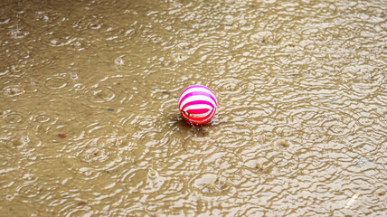 A red and white striped ball lying on a rain-soaked ground during a heavy downpour in a garden...