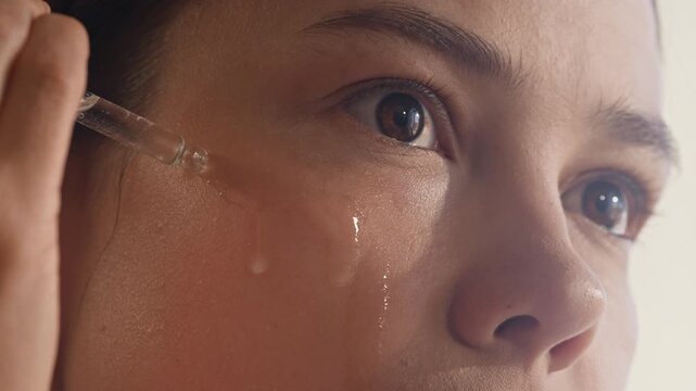 Part of face of young woman dropping rejuvenating serum on under eye zone with help of pipette while looking at herself in mirror during self care procedure