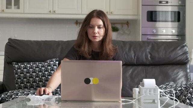 A diligent woman reviews and removes newly printed shipping labels from a label printer connected to her laptop while working from home in her kitchen.