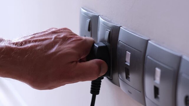 Man hand plugging a black electrical plug into a wall socket. Several sockets are aligned on a white wall in a modern interior.