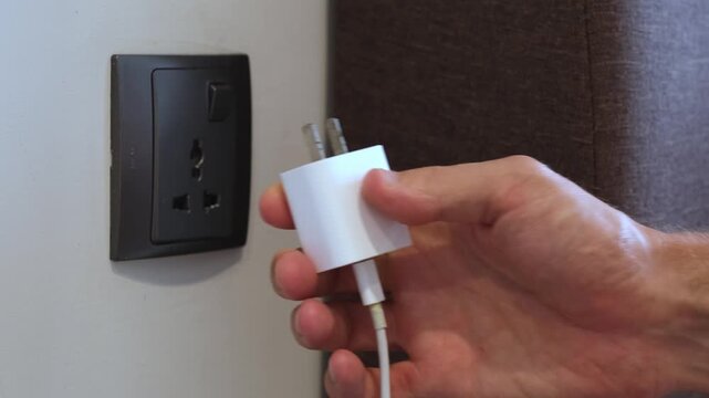 Man hand inserting a white charging adapter into a wall socket. Close-up view inside a modern interior. Technology, charging, modern lifestyle concept.