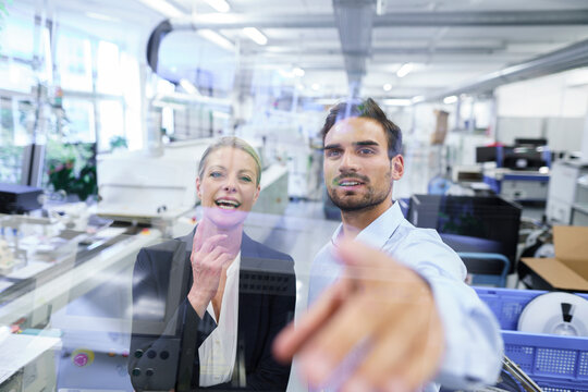 Cheerful mature businesswoman standing by businessman pointing at glass interface at factory