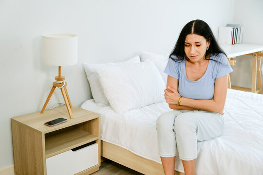 Upset woman sitting on bed, holding stomach and arms in discomfort, showing PMS symptoms and emotional stress. Concept of self-care, hormone changes, and wellness in a calm, minimal bedroom setting.