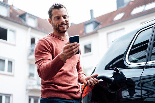 Smiling man using smart phone charging electric car outside building