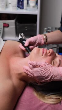 Cosmetologist performing non-invasive facial procedure on a young woman using high-tech device. Vertical video