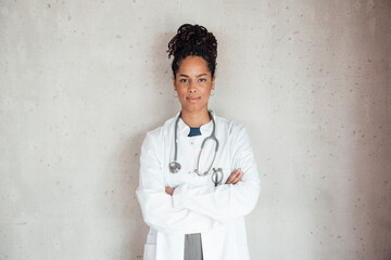 Confident young female doctor wearing lab coat with arms crossed leaning on gray wall at hospital