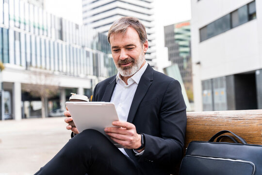 Businessman holding disposable coffee cup using tablet PC sitting on bench