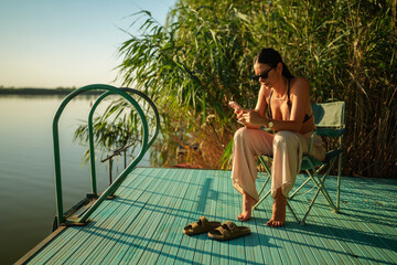 Young woman is enjoying a relaxing evening on a wooden pier by the lake, sitting on a camping chair...