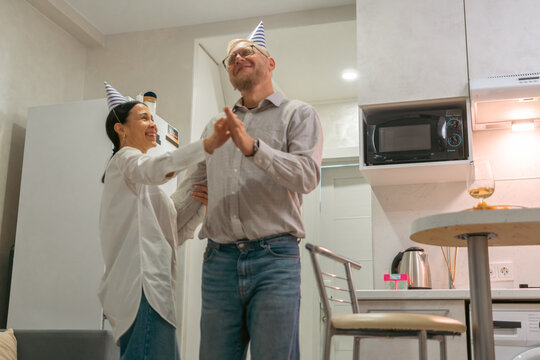 Couple Birthday Celebration Home Party: Happy mature couple joyfully dances in their home kitchen, celebrating a birthday with party hats. - Powered by Adobe