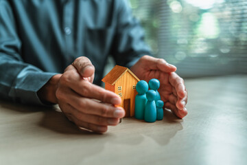 Businessman's hand gestures to protect money, house, wooden dolls represent family. Image represents the concept of asset protection, emphasizing safeguarding financial investments and property