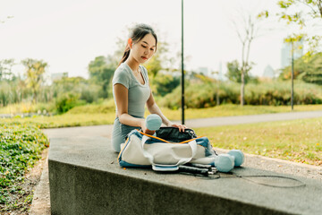 A woman in sportswear sits on a park bench, preparing for a workout with dumbbells, a jump rope, water bottle, and gym bag filled with fitness essentials.