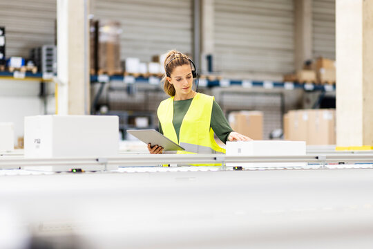 Young worker with tablet PC examining box on conveyor belt in warehouse
