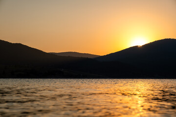 Radiant sun peeking over Rhodope mountains and reflecting off still lake water, early morning light. Morning light shimmering on surface  Dospat Lake in Bulgaria, golden reflection, calm start to day