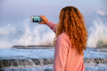Woman takes video of wave impact during golden hour, dynamic ocean background and calm posture contrast beautifully. Beach moment where red-haired woman records crashing wave, emotional and energetic 