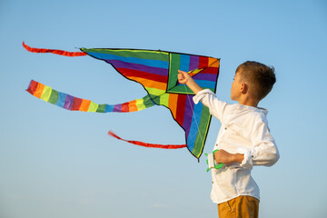 Boy holding spool with flying kite over sea, clear blue sky and water, multicolored kite soaring freely in air, capturing spirit of adventure and childhood during coastal summer break.