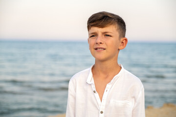 Portrait of cheerful boy in white shirt with blue sea sky in background. Serene and sunny moment from a summer holiday. Laughing child at seashore wearing a white shirt captured during golden hour.