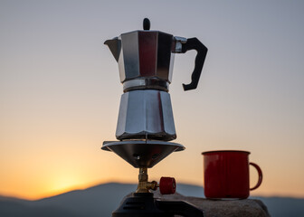 Gas stove with espresso maker on rocky shore of tranquil lake, red cup waiting at golden hour, minimalist travel concept, peaceful rest after hike, nature comfort.Scenic view of moka pot and cup lake