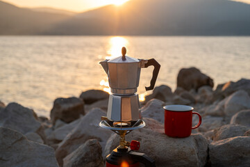 Red metal cup and moka pot on portable stove against peaceful lake and hills background at dusk, perfect scene for rustic travel, hiking break, nature lovers, coffee ritual