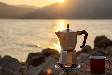 Cozy outdoor coffee setup by lake at sunset, moka pot and red mug on rocks, warm backlight over...