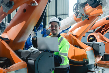 Industrial engineer giving thumbs up while inspecting large robotic arms with laptop inside advanced manufacturing facility. Image captured in natural light, highlighting confidence.