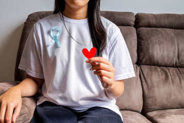 Woman Sitting On Sofa While Wearing A White Shirt With A Blue Ribbon Holding A Red Heart