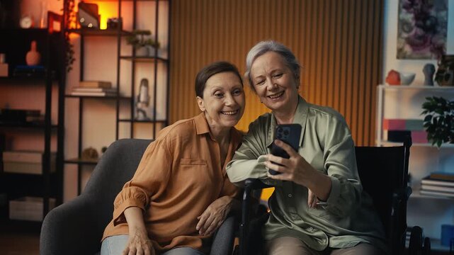 Happy senior lady with disability taking selfie on phone with her female friend