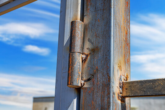 Close-up of a rusty metal hinge welded to a steel frame structure under a bright blue sky, showing signs of weathering and corrosion. - Powered by Adobe