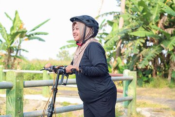 Smiling Asian young muslim woman wear hijab headscarf riding bicycle with book on basket in the park. Education and field trip outdoor concept