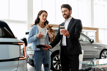 Car salesman showing credit card to woman holding charging cable for electric vehicle