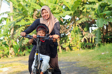 Fototapeta premium Small boy riding his bike for the first time. Young mom helping son riding a bicycle. Mom teaches her little son to ride a bicycle in the city park. Happy family moments. Time together mother and son.