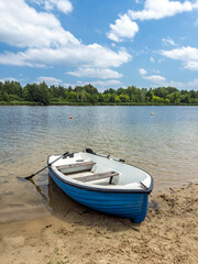 Obraz premium A blue and white rowboat with oars rests on a sandy beach by a calm lake on a beautiful sunny summer day.Peaceful summer landscape with a boat on the shore