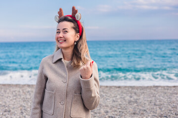 A young Caucasian woman with brown hair wears reindeer antlers at the beach during winter. The ocean is visible in the background, creating a festive atmosphere.