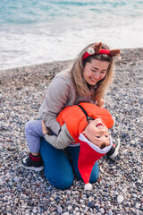 A mother and her young son play on a beach during winter. The mother has blonde hair and wears a festive headband. The son wears a Santa hat and an orange jacket.