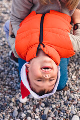 A mother holds her young son upside down at the beach during winter. The boy wears an orange puffer jacket and a Santa hat, smiling joyfully.